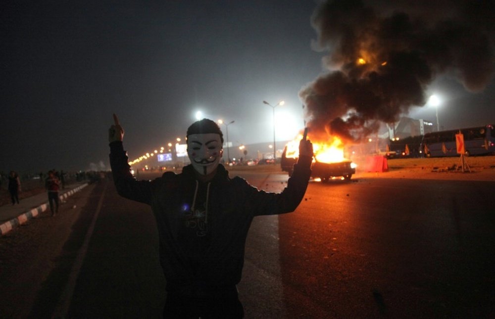 An Egyptian man wearing a mask of the anonymous movement gestures near a burning car outside a sports stadium in a Cairos northeast district, on February 8, 2015, during clashes between supporters of Zamalek football club and security forces