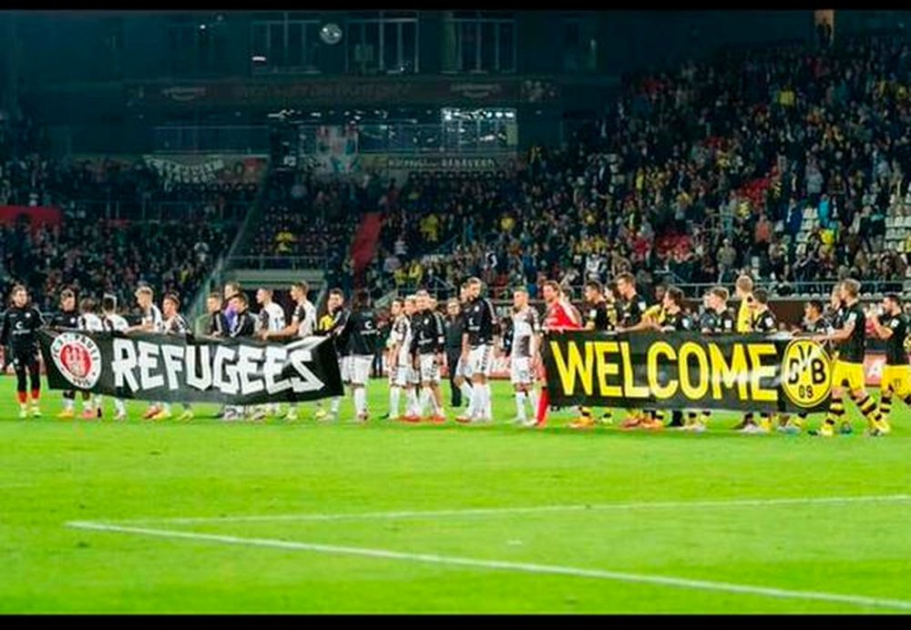 Los jugadores de St. Pauli y Borussia Dortmund sostienen unas pancartas de apoyo a los refugiados antes de iniciarse el encuentro. Twitter